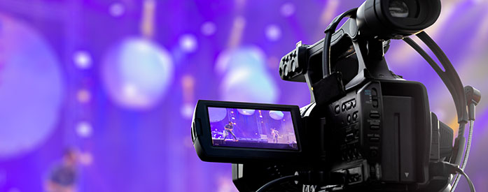 A movie camera on tripod in front of a stage with musicians playing on a stage set with purple lighting